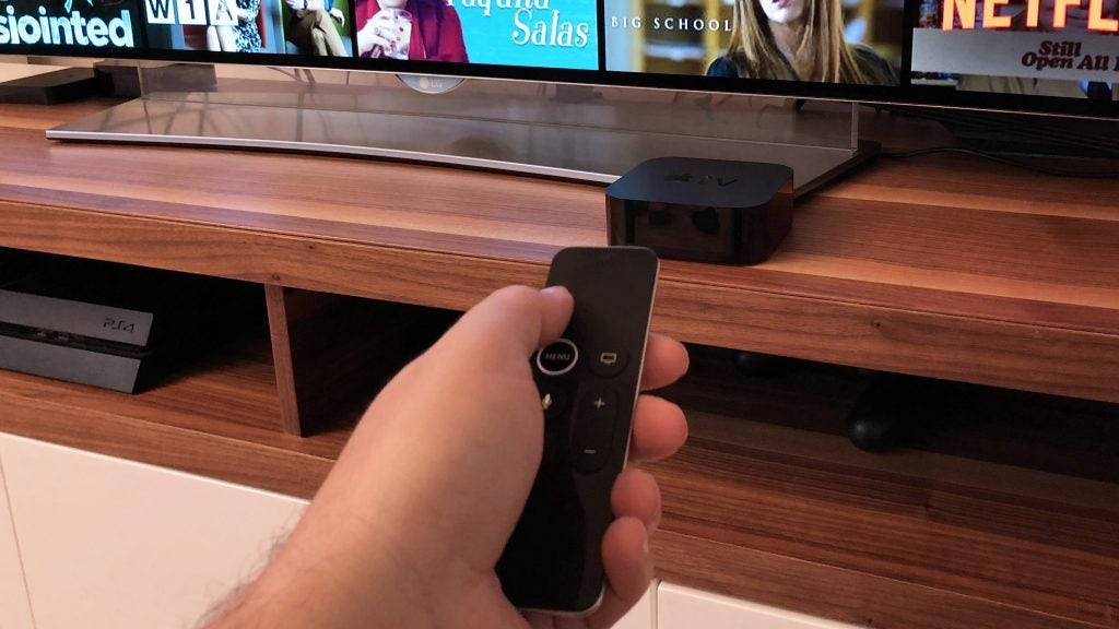 Hand holding Apple TV 4K remote with television in background.