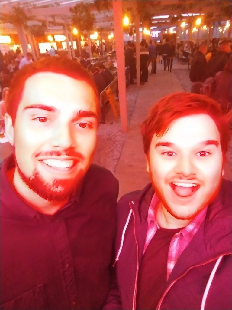 Two smiling men taking a selfie with reddish lighting at an outdoor event.