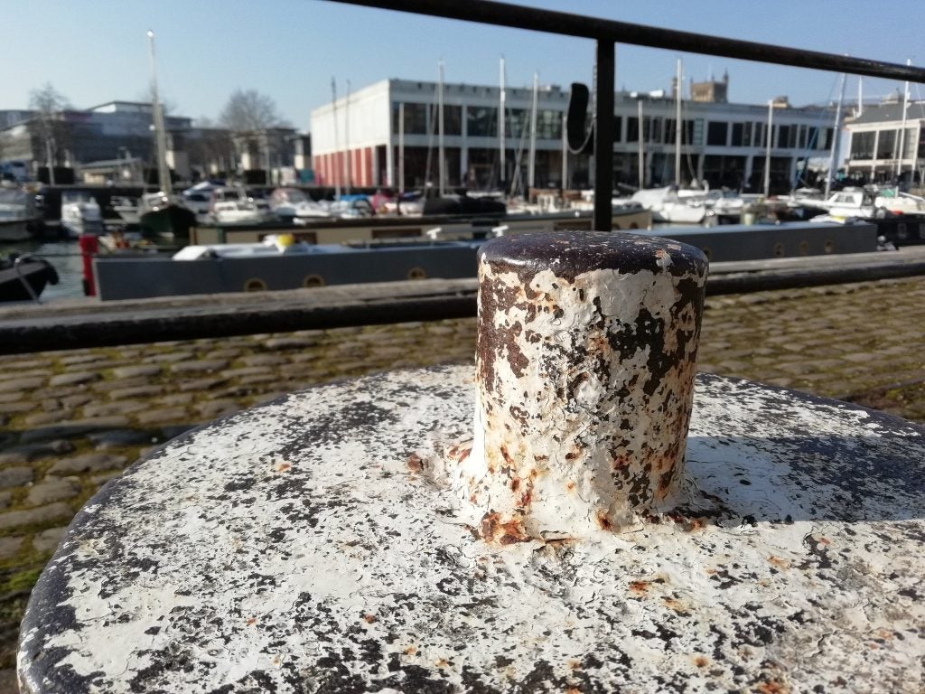 Rusty mooring bollard at sunlit marina with boats and buildings.Harbor view with historical tall ship and boats in Bristol.