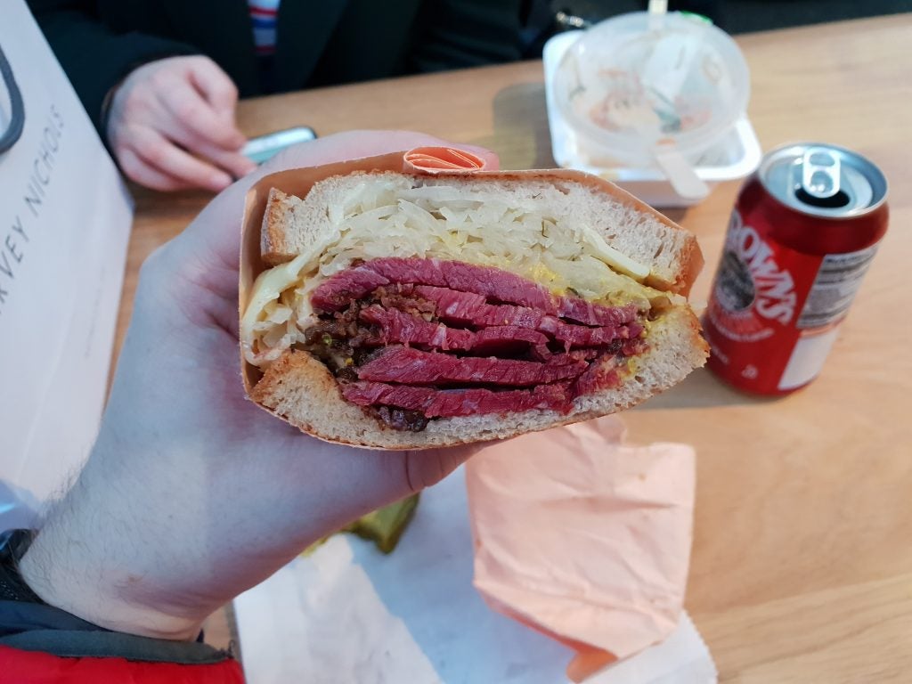 Hand holding a pastrami sandwich with soda can in background.