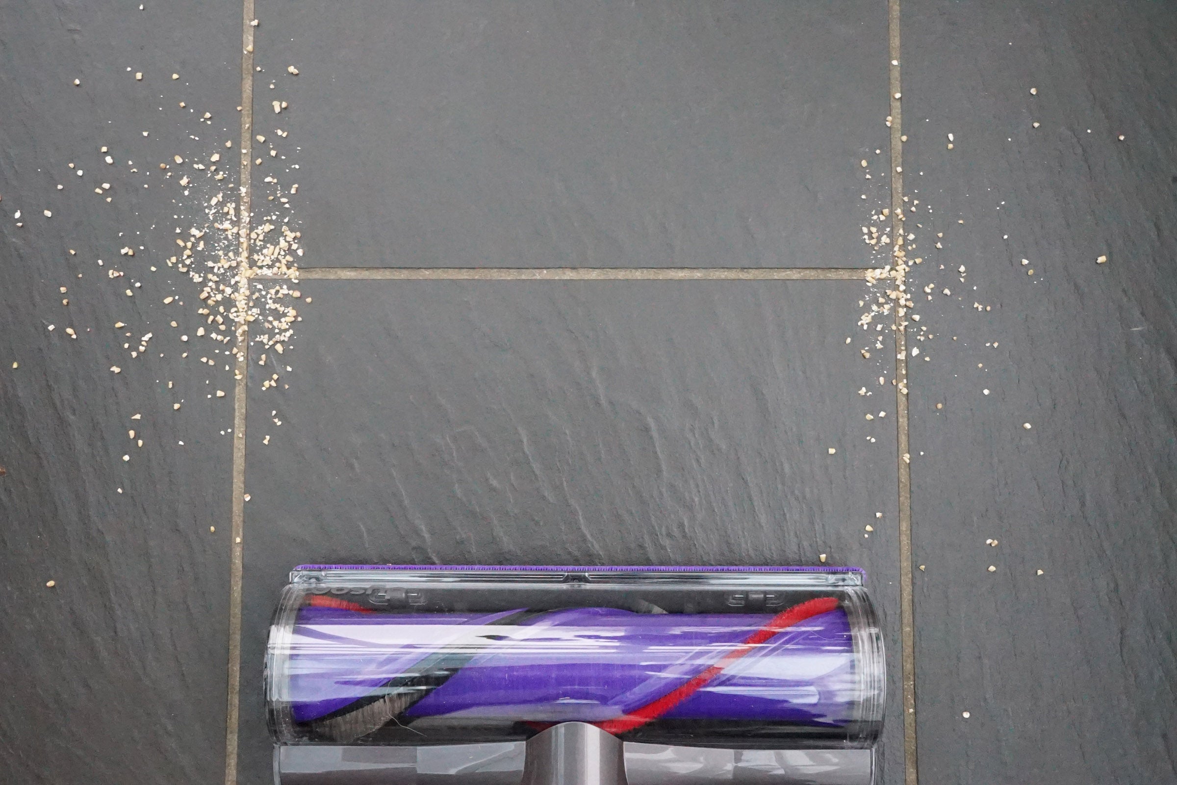 Dyson Cyclone V10 vacuum cleaning oats off dark tile floor.