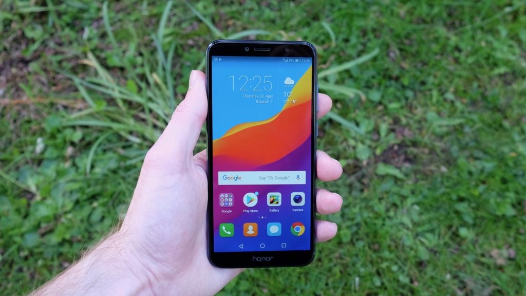 Hand holding Honor 7A smartphone displaying home screen outdoors.