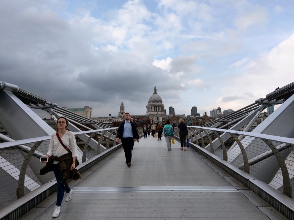 Pedestrians on a bridge with cloudy sky and cityscape.