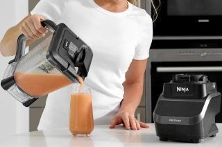 A black Ninja CT610UK blender standing on table with it's jar held in hand pouring drink in glass