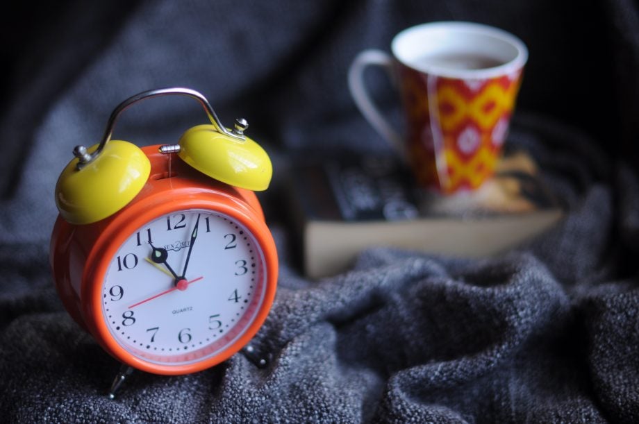 Close up view of an alarm clock kept on bed with a mug and a book kept beside