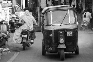 A black & white picture of a rickshaw on road with shops behind and other vehicles around