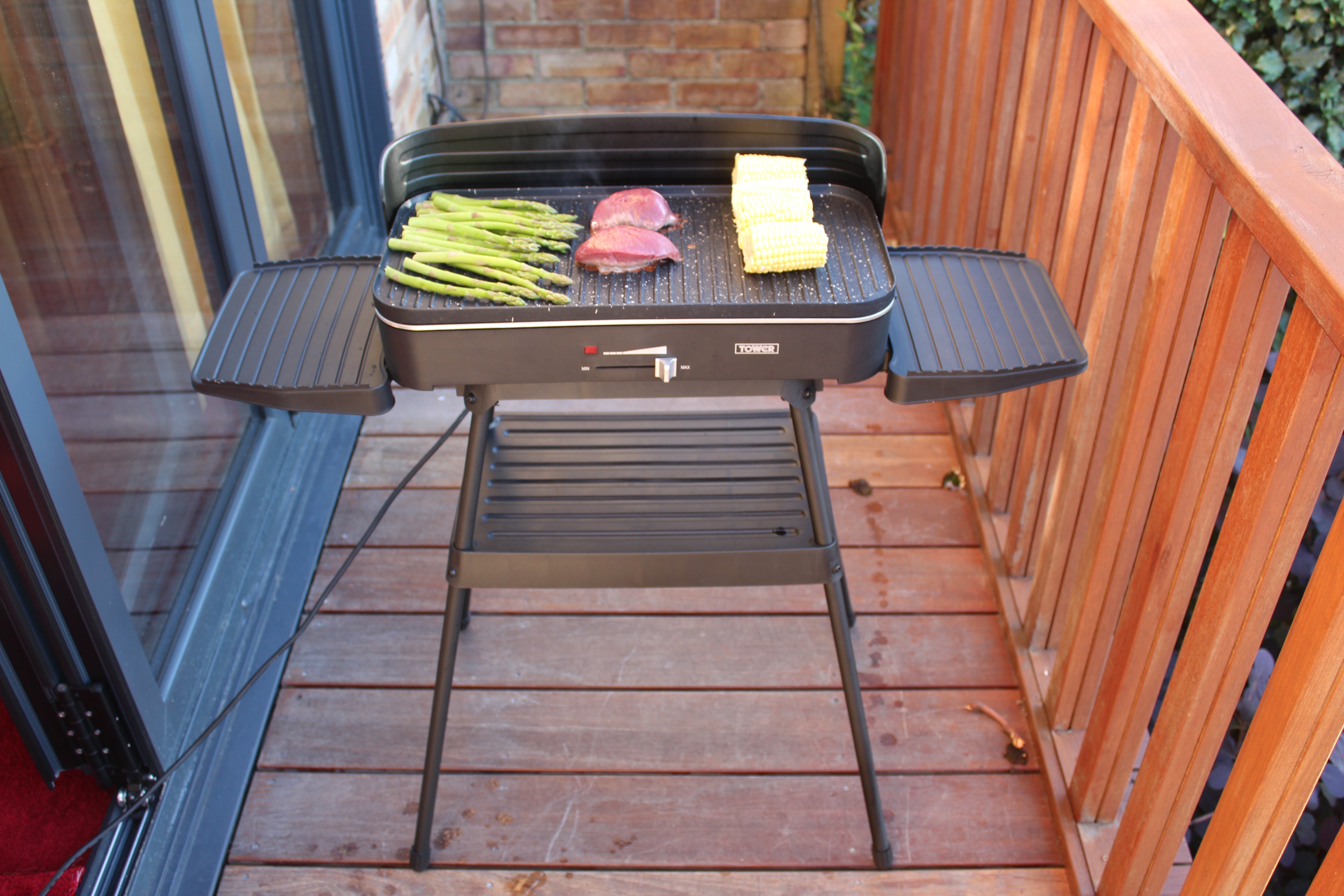 Front top view of a black Tower T14028 with stand standing on floor with different food items being grilled on it