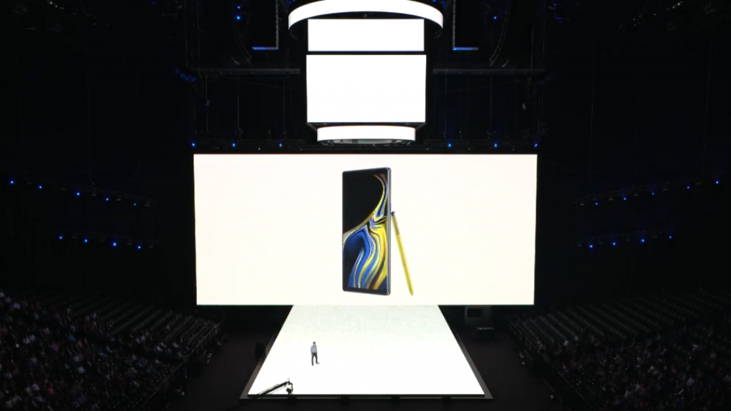 A man standing on stage with Samsung Galaxy Note 9 with it’s S-Pen leaning beside displayed on screen behind