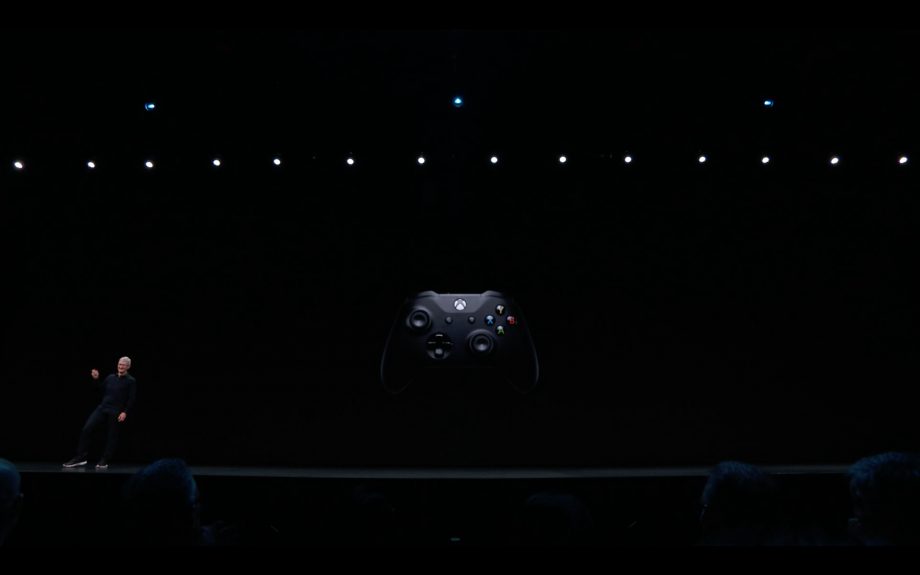 A man standing on a stage with a black Xbox gaming controller displayed on the screen behind