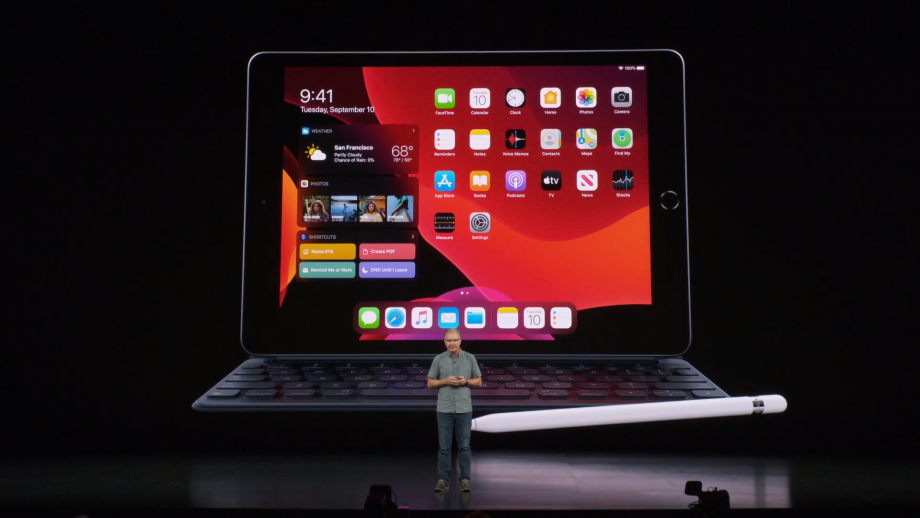 A man standing on stage with an iPad 7th Gen with keyboard attached and it's pencil kept on it displayed on screen behind