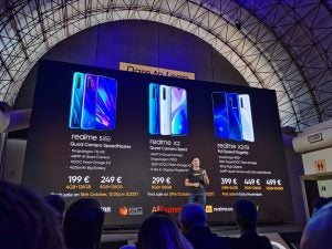 A man standing on stage facing audience with Realme 5 Pro, X2 and X2 Pro displayed on screen behind
