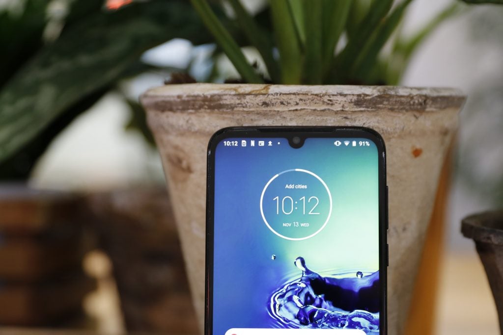 A Motorola G8 Plus standing against a plant pot displaying homescreen, top half close up view