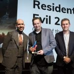 A man receiving award from two men on stage, Resident Evil 2 displayed on screen behind, Trusted Reviews Awards