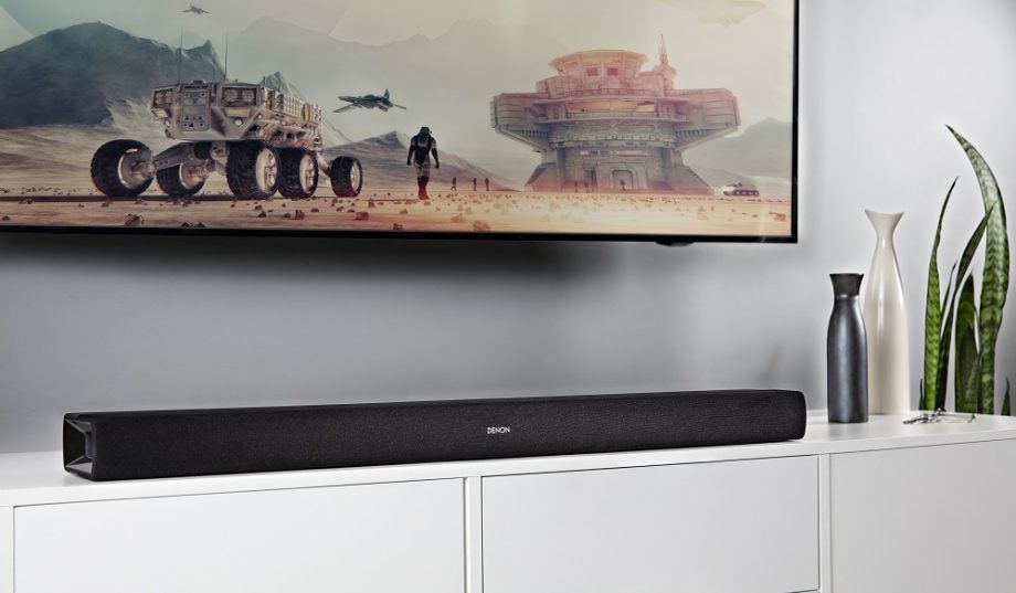 A black Denon DTH 216 Studio soudbar kept on a table below a mounted TV on wall