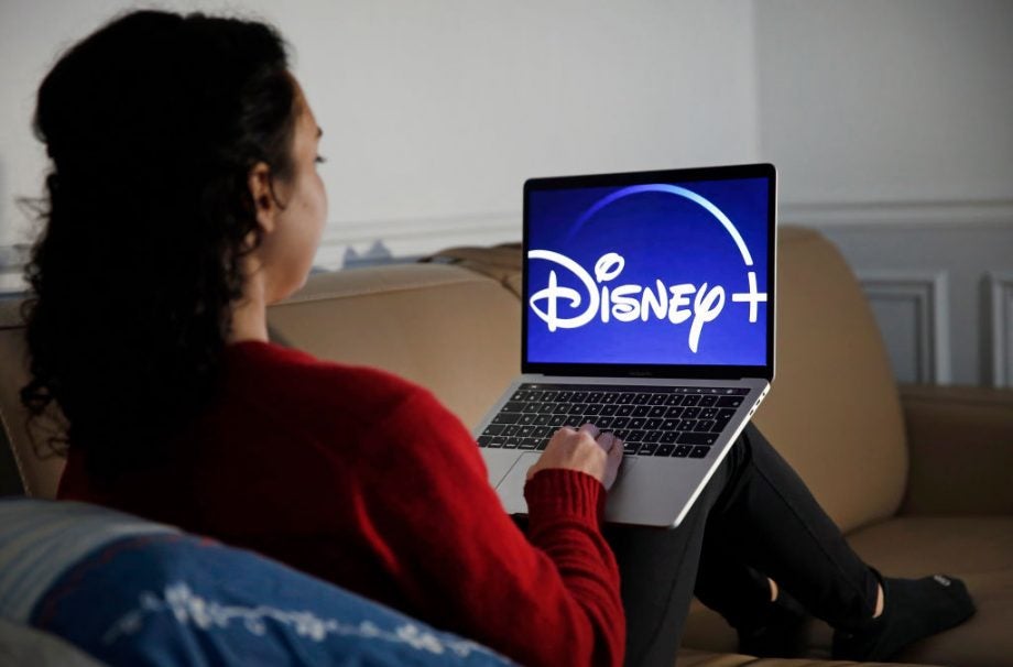 A woman in red-black outfit sitting on a couch using a Macbook Pro displaying Disney+ wallpaper