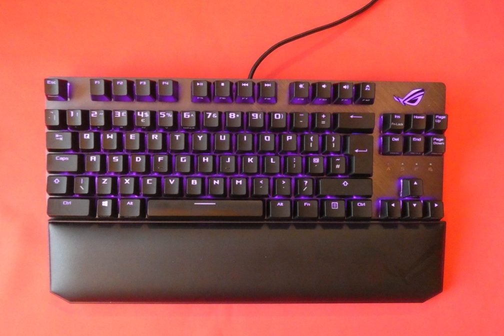 ASUS ROG Strix Scope TKL DeluxeA black ROG Strix Scope TKL Deluxe keyboard kept on a red background with it's wrist rest attached and purple lights beneathe the keys