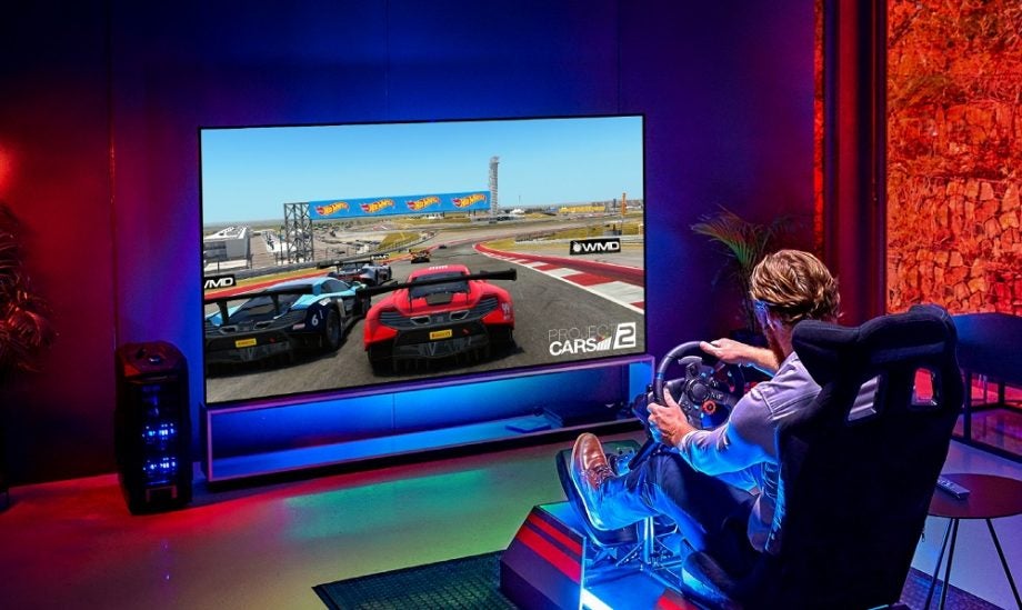 A black LG OLED ZX TV standing on a shelf displaying a car race being controlled by a man sitting on simulator