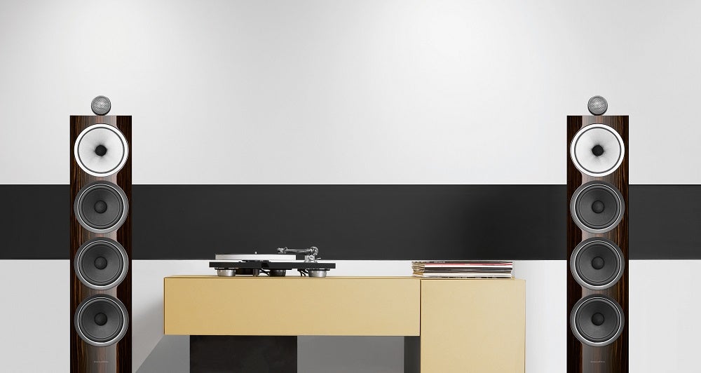 Two brown-black BW702 signature speakers standing in a room on either sides of a table