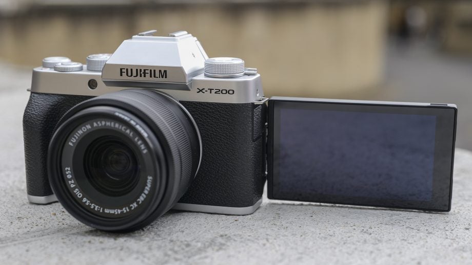 Front panel view of a silver-black Fujifilm XT200 standing on a concrete floor with its back screen flipped on side facing front