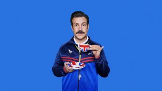 Ted Lasso standing on a blue background in coach outfit drinking tea