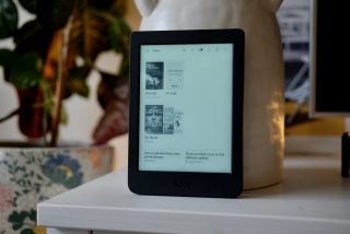 A black Kobo e-reader standing on a white table displaying homescreen with books