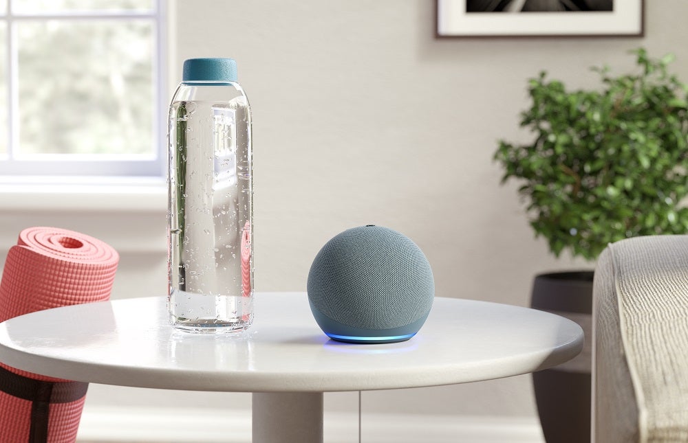 A silver-blue Amazon Echo Dot standing on a white table beside a water bottle