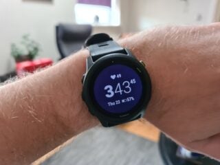 A black Garmin watch tied on wrist displaying date, battery and heart details