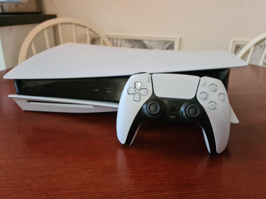 A PS5 laid on a wooden table with it's controller standing against it