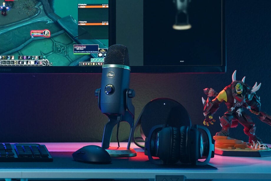 A Blue micrpohone standing on a table with headphones, keyboard, mouse and monitor kept around