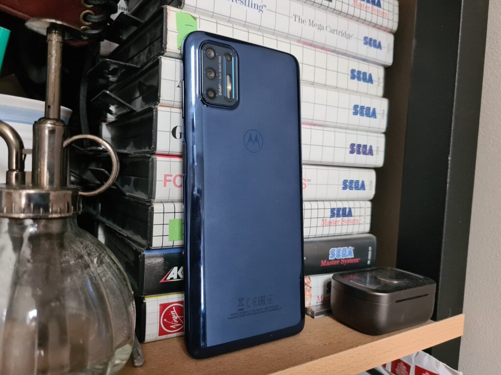 Back panel view of a blue Motorola G9 Plus standing against a shelf made of books