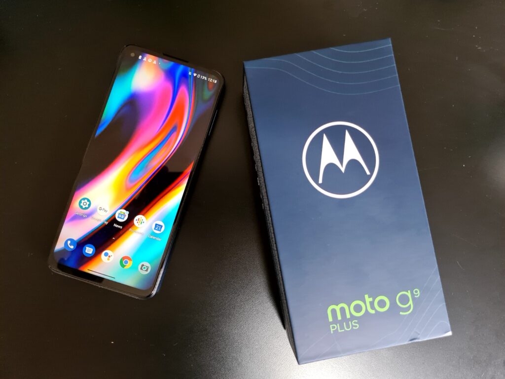 View from top, of a black Moto G9 Plus resting on a black table with its box beside