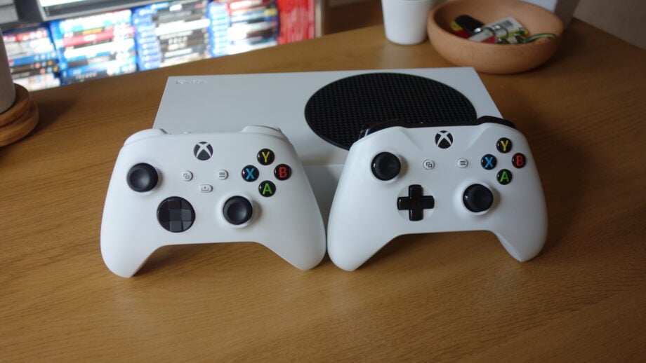 Two white Xbox controllers standing against a white Xbox on a wooden table