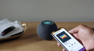 A black Homepod mini standing on a wooden table and smartphone playing music held in hand beside