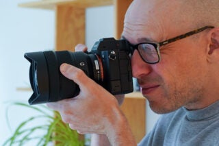 A man wearing glasses holding a black Sony A7s iii camera to click a picture