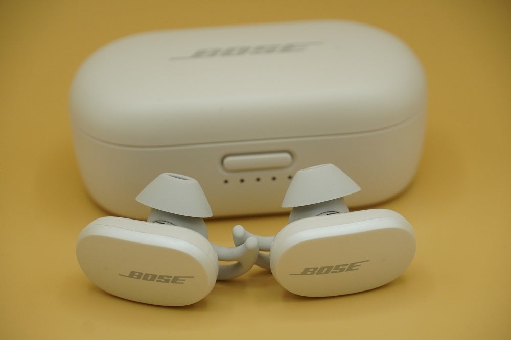 White Bose QuietComfort earbuds resting on yellow background with it's white case standing behind
