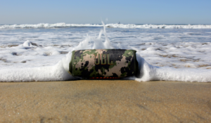 A green-black JBL Charge 5 speaker resting on shore