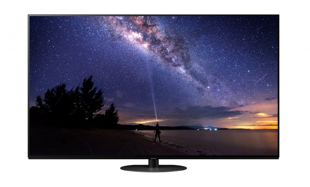 A black Panasonic JZ1000 TV standing on a white background displaying a man standing on shore pointing a torch towards the sky