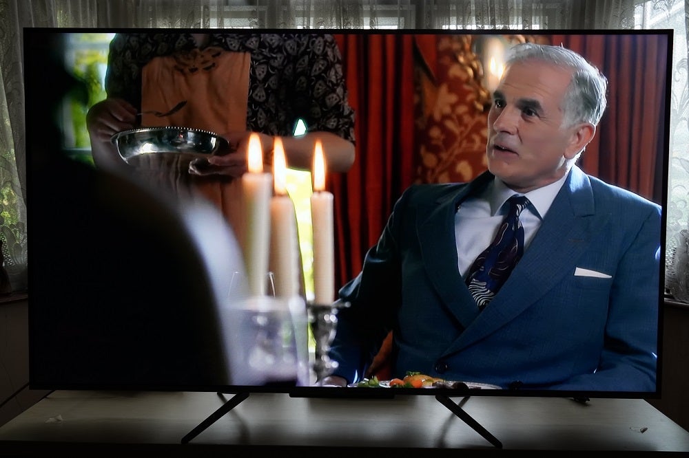 A black TCL-55C715K TV standing on table, displaying an old man sitting at a dinner table