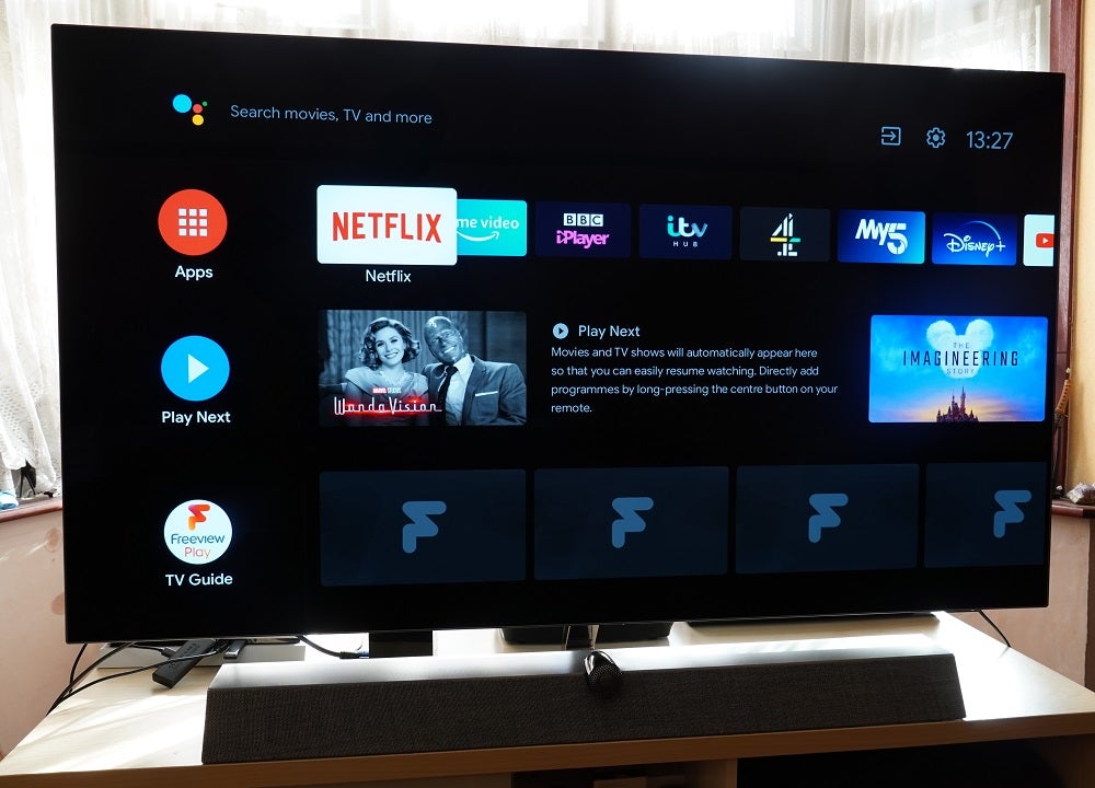 A black Philips OLED935 standing on a table, displaying home screen with apps, play next and TV guide sections
