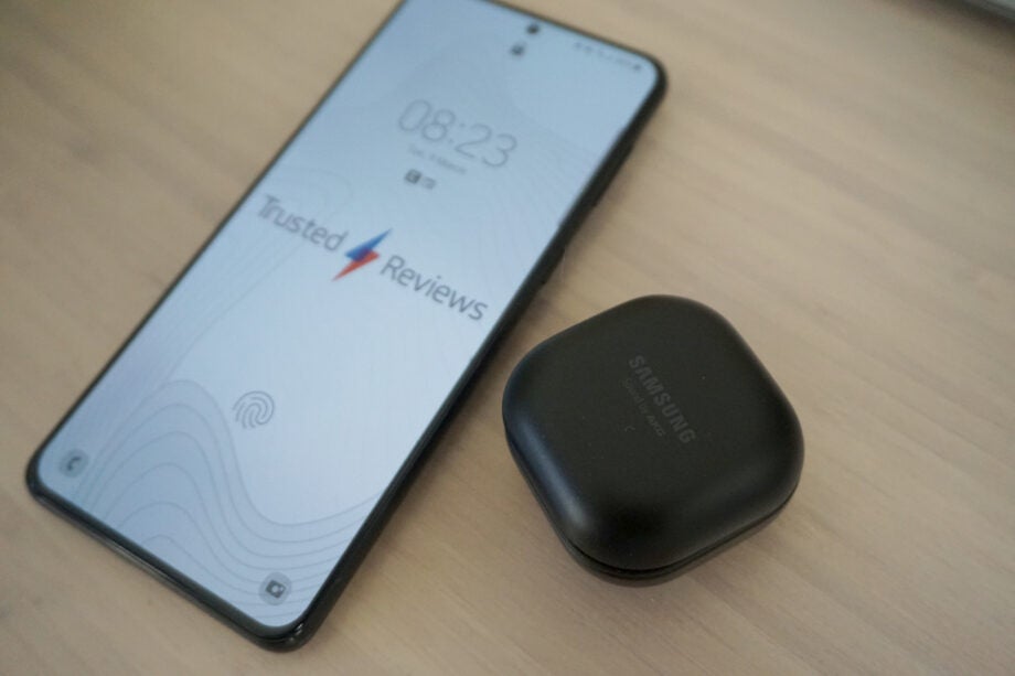 A smartphone displaying Trusted reviews logo on lock screen laid on a table and a black case of Galaxy buds pro resting beside