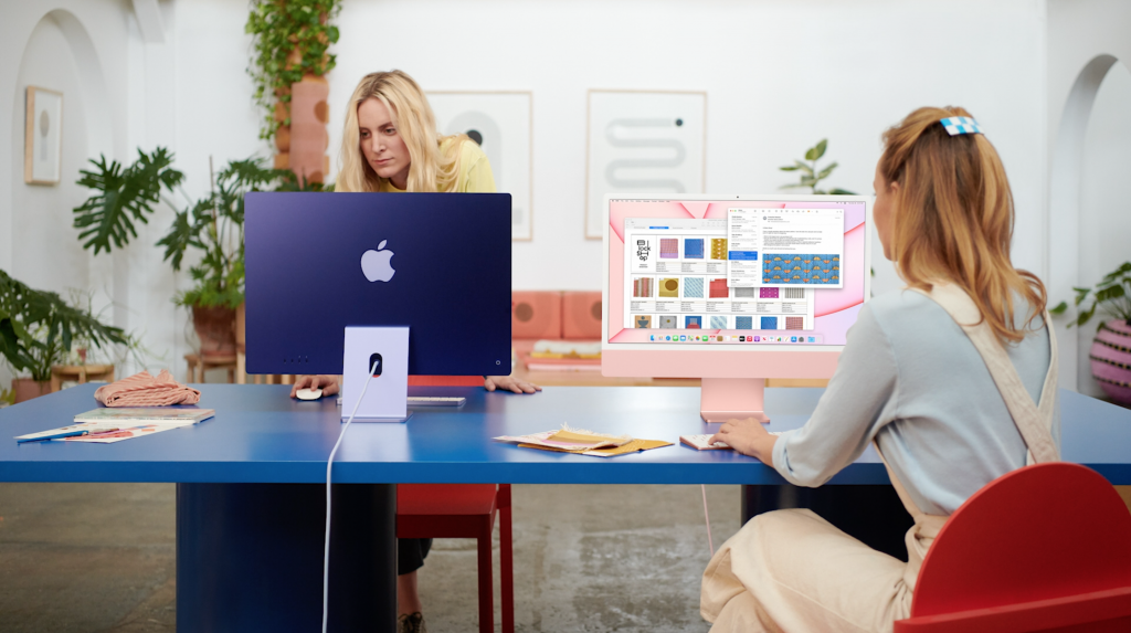 Two women sitting on either side of a blue table, using iMacs of blue and light pink color