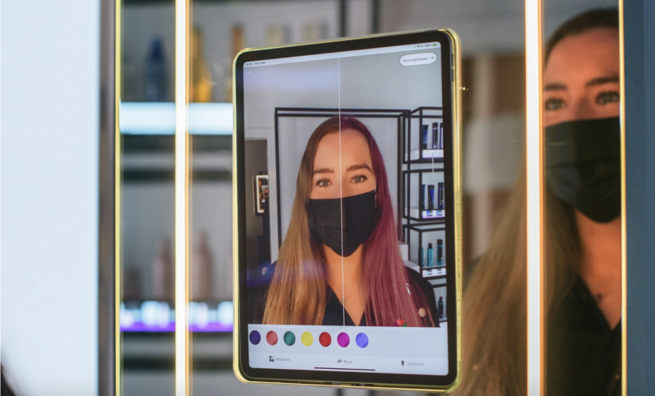 A tablet attached to a mirror with a software displayed to check you’re your hair color in different colors, a blonde girl looking in the tab, checking how she would look in burgndy colored hairs