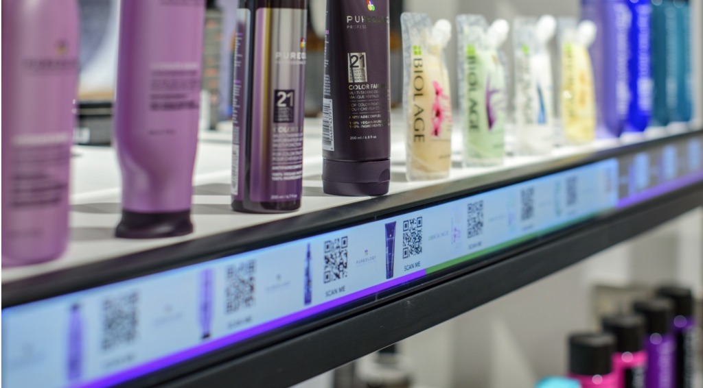 Close up image of various cosmetic tubes and bottles standing on a rack with their barcodes printed below on the rack's front edgeA tablet attached to a mirror with a software displayed to check you’re your hair color in different colors, a blonde girl looking in the tab, checking how she would look in burgndy colored hairs