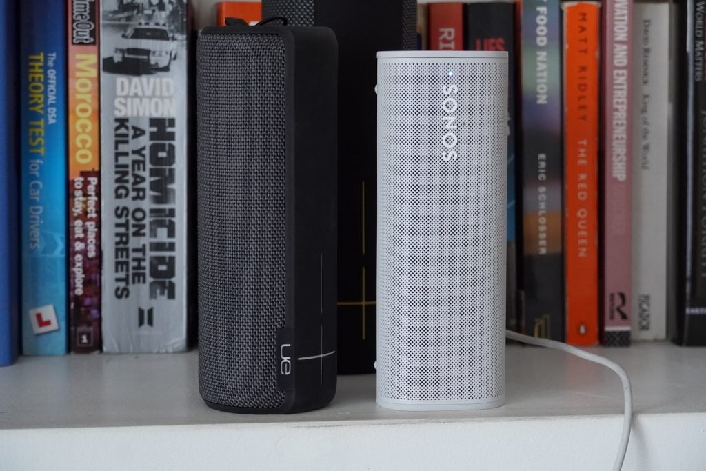 A black UE Boom 2 speaker standing beside a white Sonos Roam speaker, on a book shelf's edge