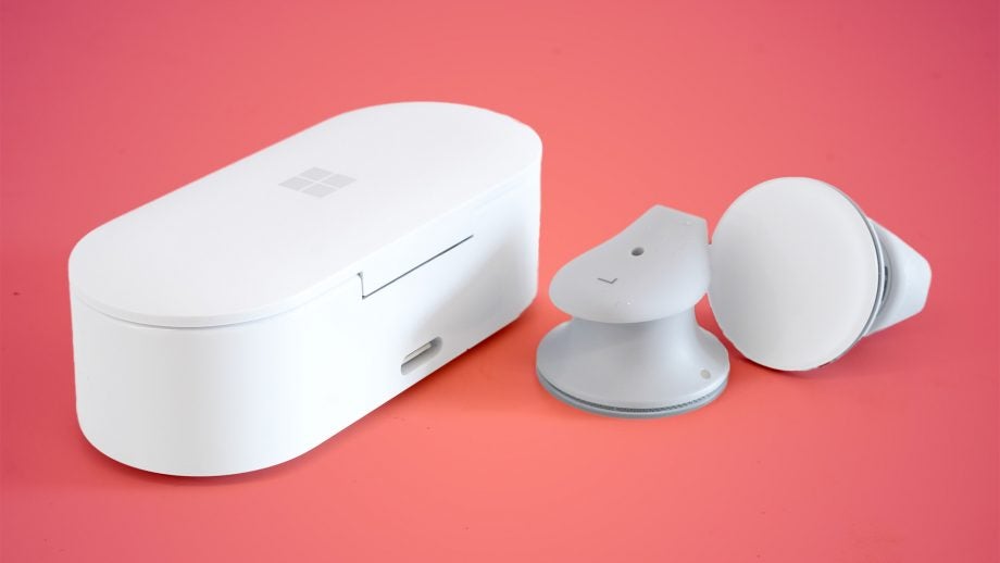White closed case of Surface earbuds standing on left and earbuds resting beside on a pale pink background
