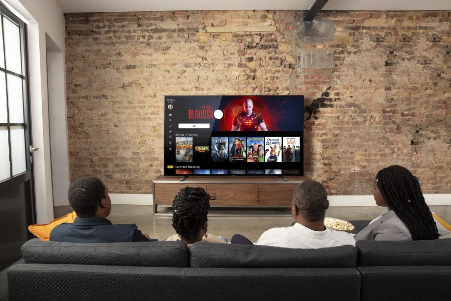 Four people, two men and women sitting on a dark grey couch facing back, watching Netflix on Bravia A80J core standing on a wooden shelf
