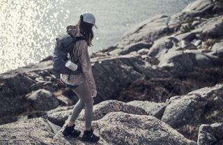 A girl with in hiking outfit and a black hiking backpack with white BO Beosound explore speaker hanging on the right, walking on a hill with a lake or river on the left