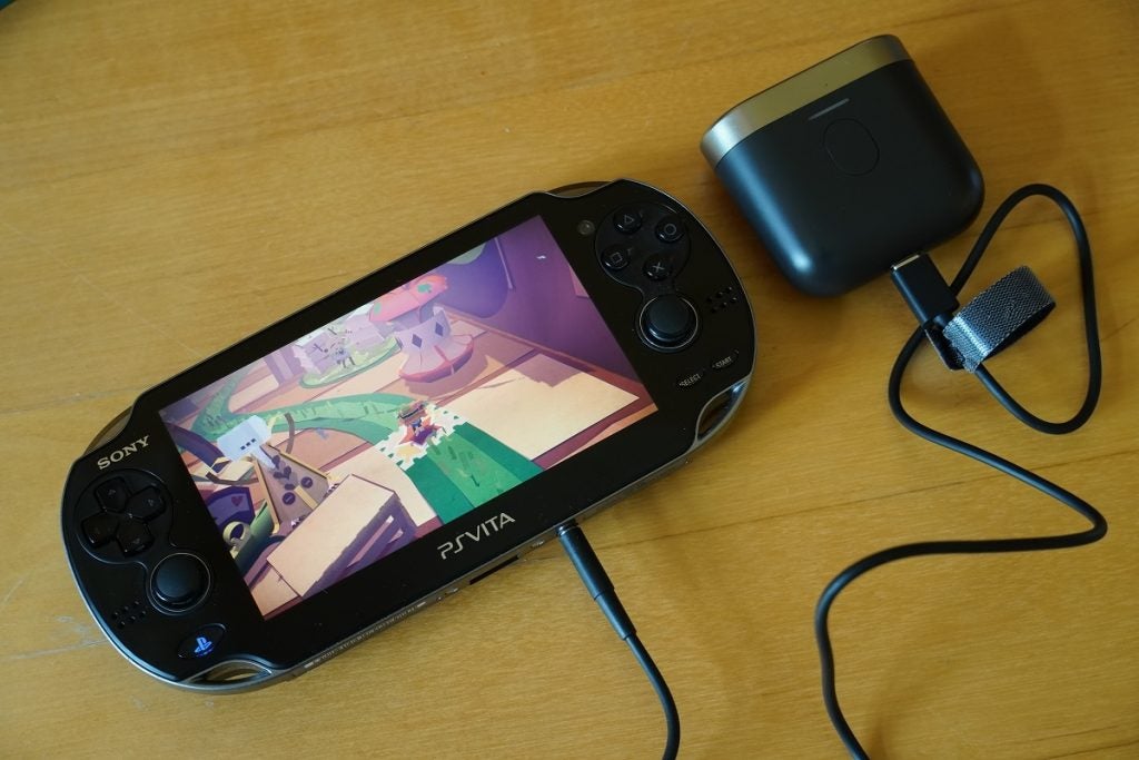 A black Sony playstation PS Vita displaying a game with gaming controls on either side, a jack connected to it and other end connected to a speaker, showcasing Bowers and Wilkins P17 audio transmission