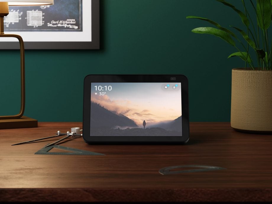 Amazon echoshow 8 of black color with black border on the screen and a wallpaper of mountains at dawn with time and temprature on top of it a plant pot with plant on the right, and scales around it on a wooden table