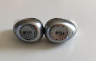Silver and white KEF Mu3 earbuds resting on white background, back side view, a video camera logo with KEF printed in the back